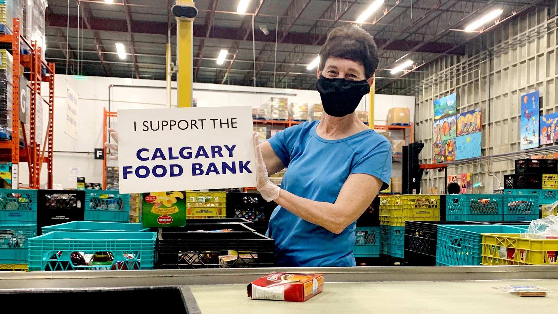 A person wearing a mask and blue t-shirt is holding a sign that says I support the Calgary Food Bank. She's standing at the sorting line at the Calgary Food Bank.